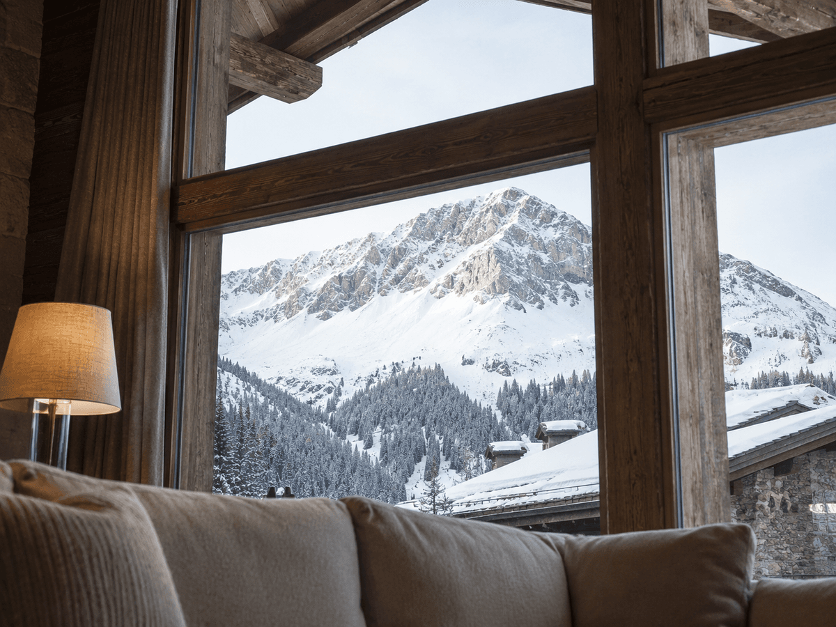 Snow-covered Alpine peaks and a chalet rooftop seen through a timber-framed chalet window, with a lamp and sofa in the foreground